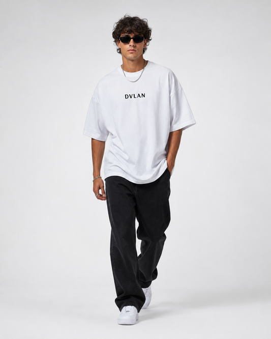 A man wearing a white oversized t-shirt with the brand name 'DVLAN' printed in black on the chest, paired with black pants and white sneakers, walking against a light gray studio background.