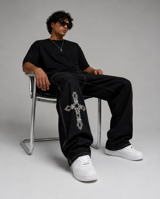 A man wearing a black t-shirt, black jeans with a large silver cross embellishment on the left leg, and white sneakers, sitting on a chrome chair against a gray studio background.