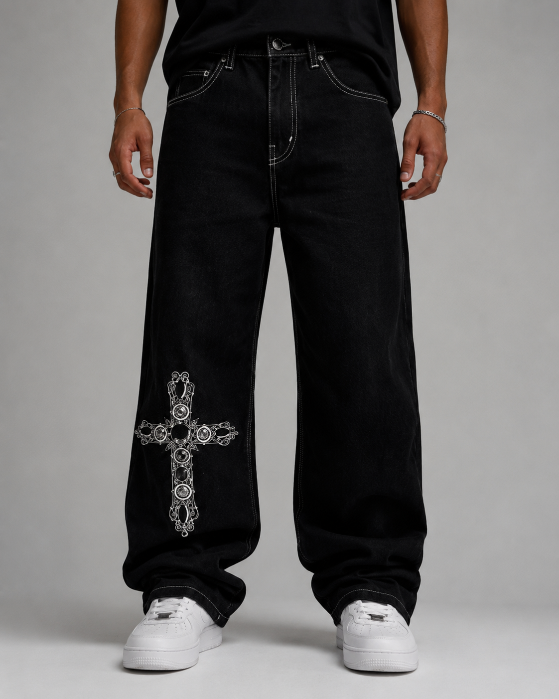 A person wearing very baggy black jeans with a large white ornate cross graphic on the lower left leg, paired with a black top and white sneakers, photographed against a light gray studio background.