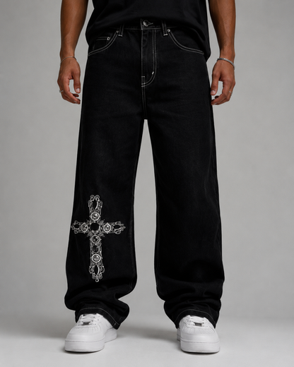A person wearing very baggy black jeans with a large white ornate cross graphic on the lower left leg, paired with a black top and white sneakers, photographed against a light gray studio background.