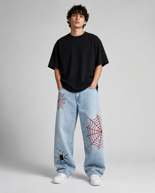 A model stands against a light gray studio background wearing a black oversized t-shirt, light-wash blue jeans with red spiderweb prints and a black spider graphic, and white sneakers.