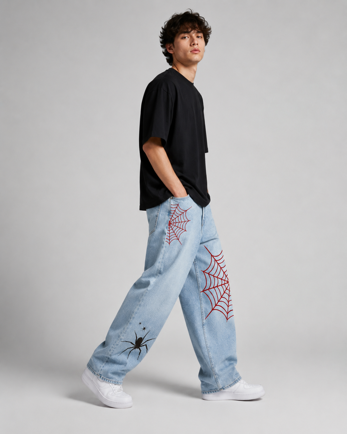 A model wearing light blue jeans with red embroidered spiderwebs and black spider graphics, paired with a black t-shirt and white sneakers, standing against a light gray studio background.