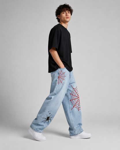 A model wearing light blue jeans with red embroidered spiderwebs and black spider graphics, paired with a black t-shirt and white sneakers, standing against a light gray studio background.