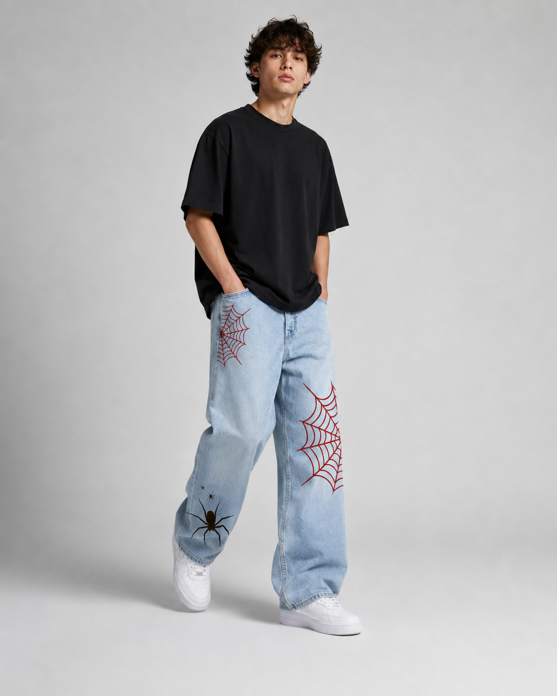 A model stands against a light gray studio background wearing a black oversized t-shirt, light-wash blue jeans with red spiderweb embroidery, and white sneakers.
