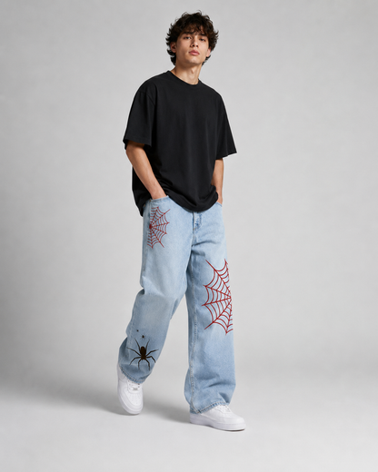 A model stands against a light gray studio background wearing a black oversized t-shirt, light-wash blue jeans with red spiderweb embroidery, and white sneakers.
