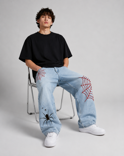 A model sits on a metal folding chair wearing light-wash blue jeans with red spiderweb graphics on the knees and a black t-shirt, paired with white sneakers, against a light gray studio background.