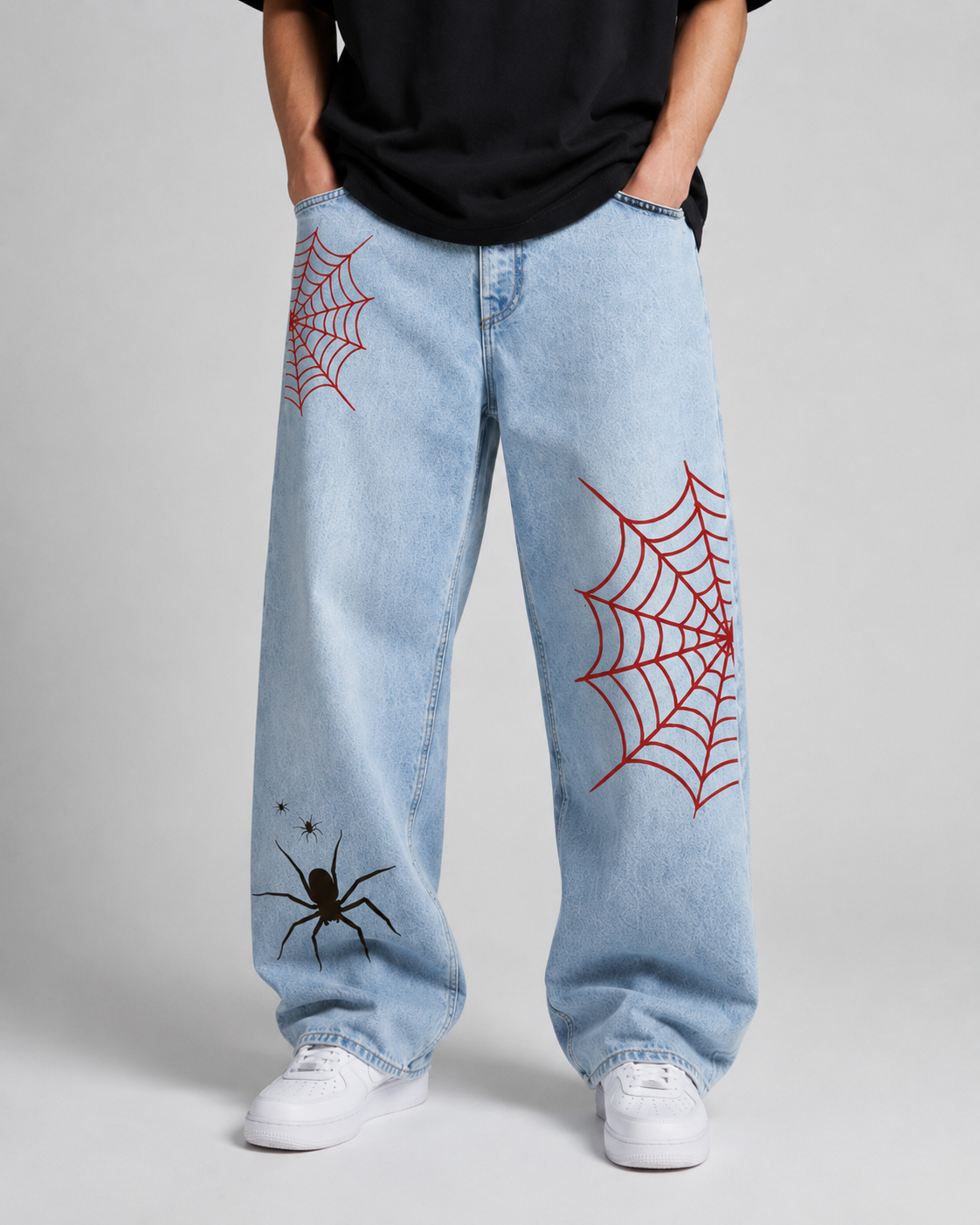 A person wearing light-wash blue jeans with red spiderweb prints and a black spider graphic on the lower left leg, paired with a black t-shirt and white sneakers against a light gray studio background.