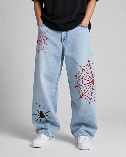 A person wearing light-wash blue jeans with red spiderweb prints and a black spider graphic on the lower left leg, paired with a black t-shirt and white sneakers against a light gray studio background.