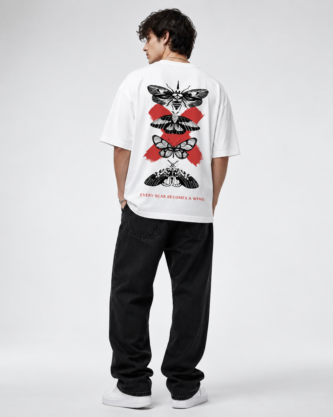 A man viewed from behind wearing a white t-shirt with a large red and black moth graphic on the back, paired with black jeans and white sneakers, against a light gray studio background.