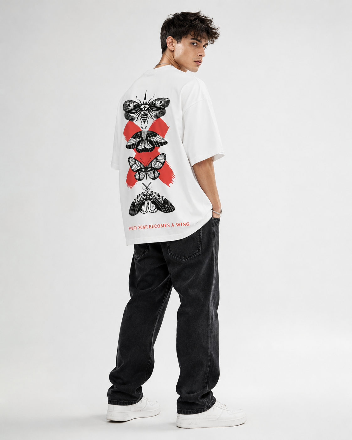 A model wearing a white oversized t-shirt with a large graphic print of stacked moths and red abstract shapes on the back, paired with black jeans and white sneakers, photographed against a light gray studio background.