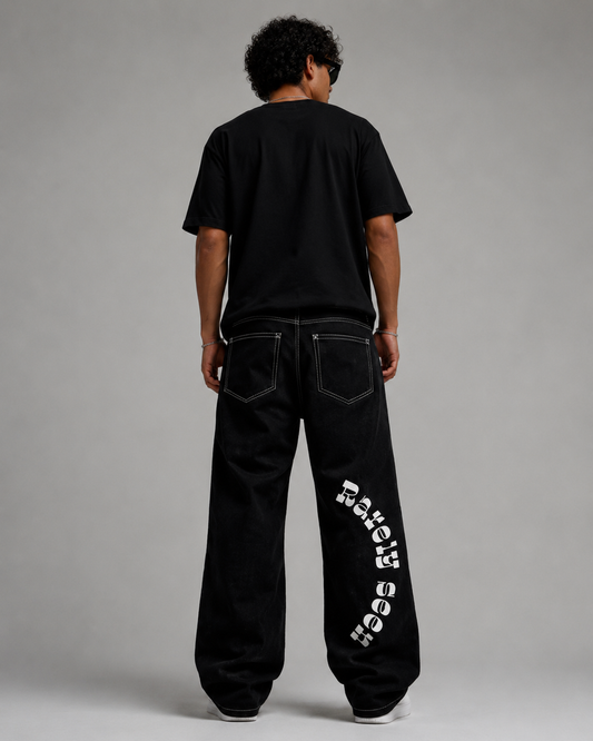 A person shown from the back wearing black wide-leg jeans with a large white 'Macy's' logo printed vertically down one leg, paired with a black t-shirt, against a neutral gray studio background.