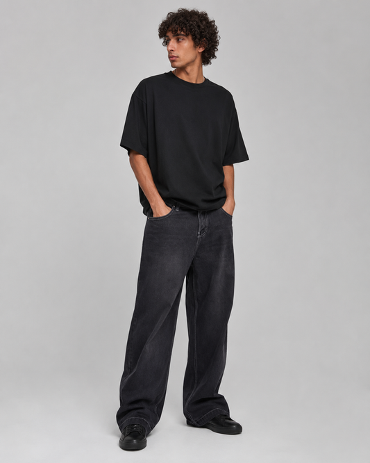 A man modeling a black oversized short-sleeve t-shirt and dark gray wide-leg jeans against a light gray studio background.