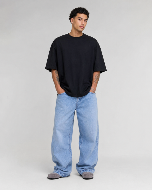 A man modeling an oversized black short-sleeve t-shirt and light-wash wide-leg blue jeans against a plain light gray studio background.