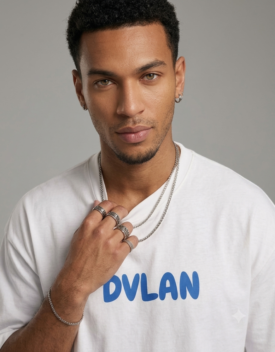 A man wearing a white t-shirt with the name 'DVLAN' printed in blue across the chest, accessorized with silver chain necklaces, rings, and a bracelet, against a plain gray background.